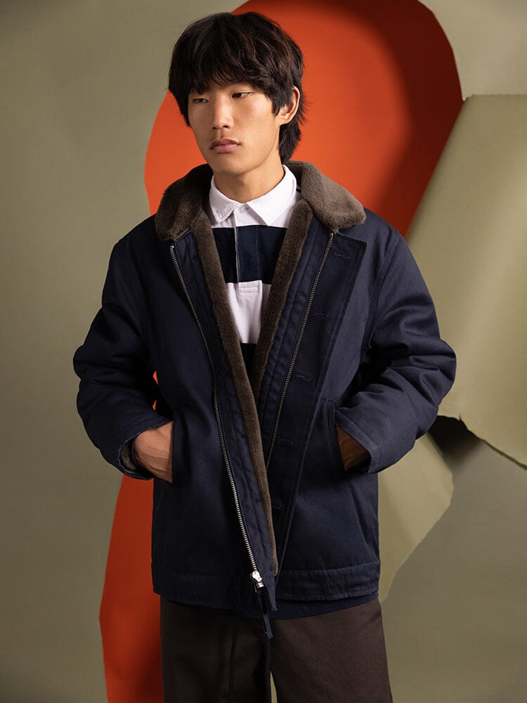A person wearing an 1940s N-1 Deck Jacket in navy blue by Alpha Industries with a fur collar stands with hands in pockets.