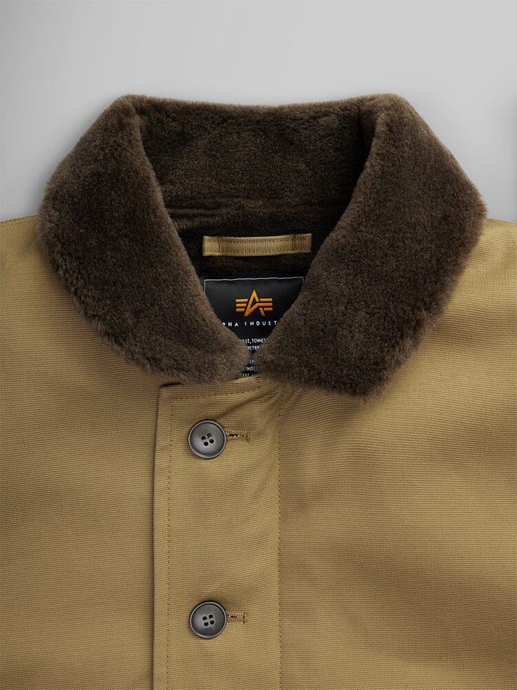 Close-up of the 1940s N-1 Deck Jacket by Alpha Industries is a classic tan deck jacket with a cotton shell. It features a dark brown fur collar, two buttons, and has a visible tag on the inside.