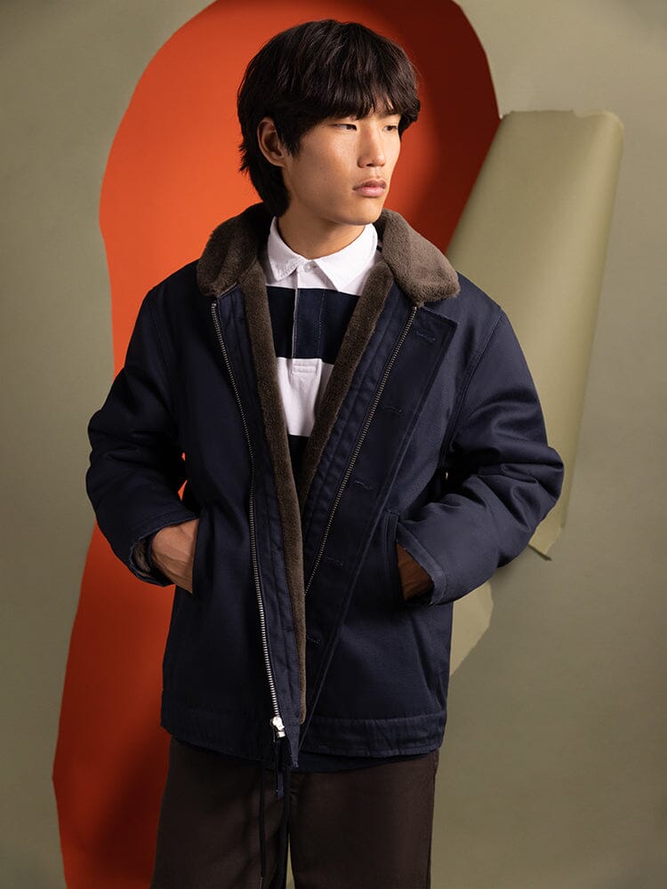 A person wearing an Alpha Industries 1940s N-1 Deck Jacket in navy blue, featuring a fur collar, gazes to the right.