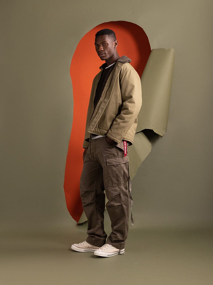 Clad in an Alpha Industries 1940s N-1 Deck Jacket in khaki, a person stands channeling the rugged style reminiscent of classic US Navy attire.