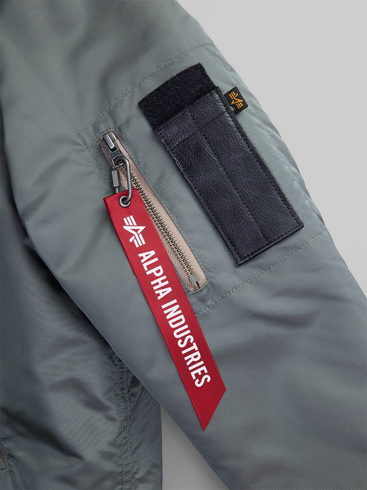Close-up of the 1960s RCAF Canadian Airforce Type III Jacket by Alpha Industries features a gray jacket sleeve with a zippered pocket, black hook and loop fastener patch, and a red tag labeled Alpha Industries.