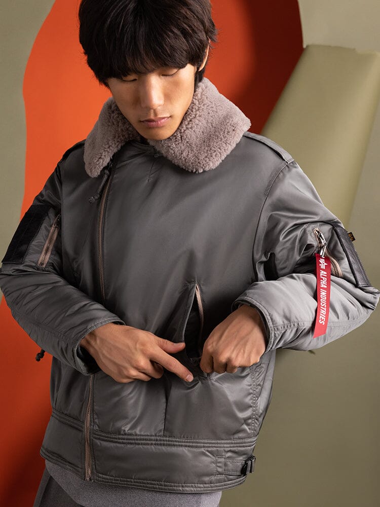 A person wearing an Alpha Industries 1960s RCAF Canadian Airforce Type III Jacket, featuring a gray design with a fur collar, adjusts the utility pocket.