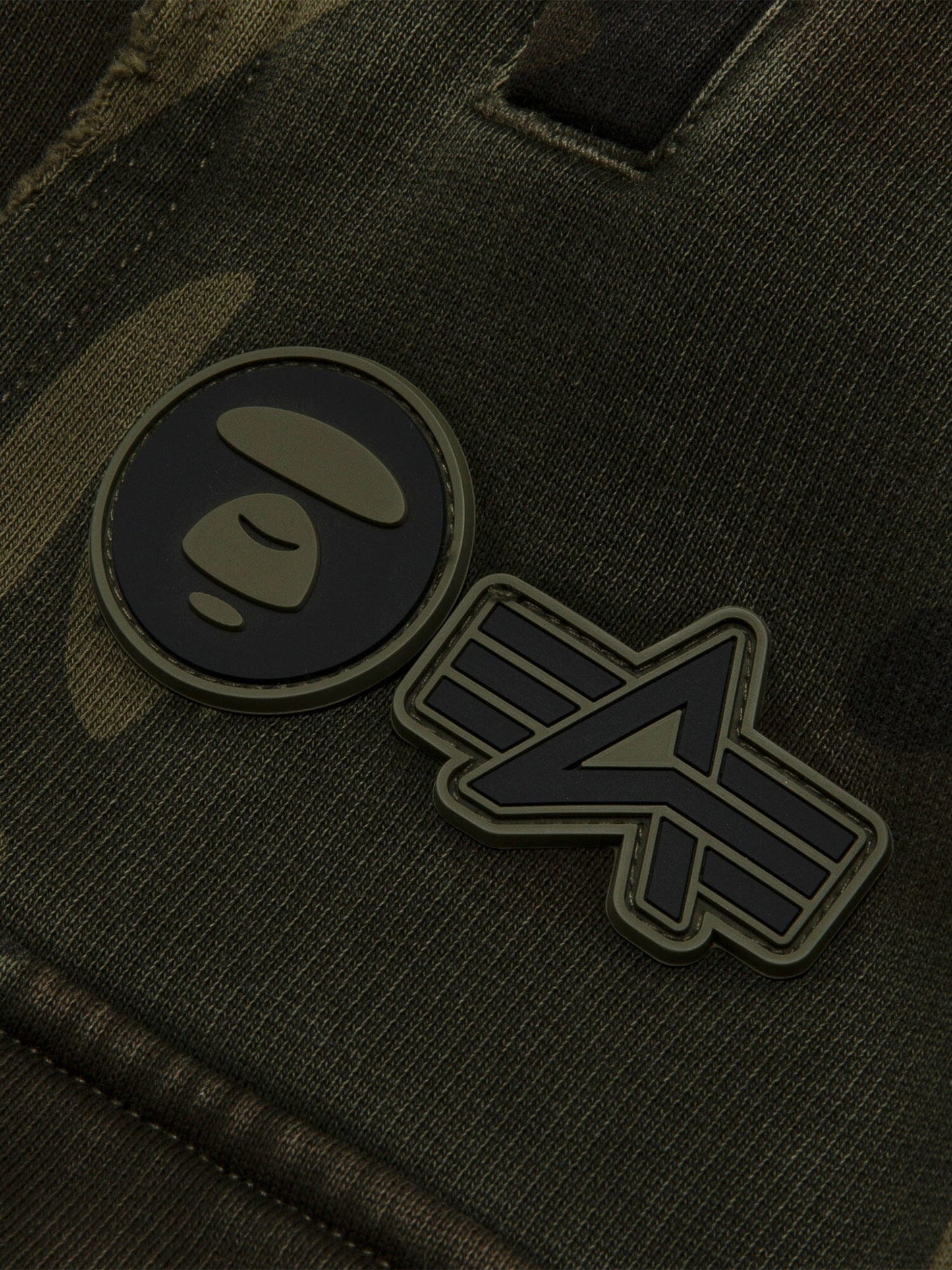 Aape X Alpha Camo Army Sweatpant by Alpha Industries, camo fabric, two rubber patches: circular face and abstract emblem.
