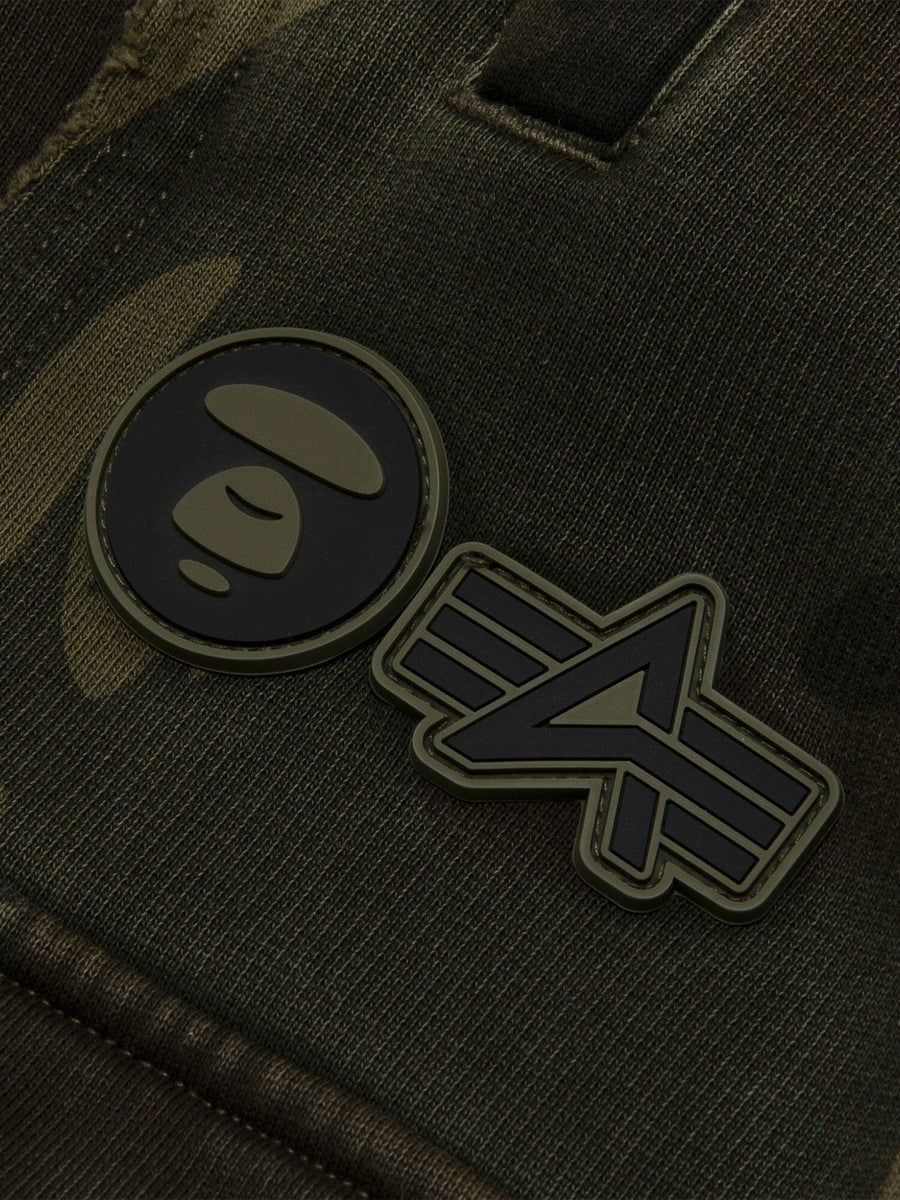 Aape X Alpha Camo Army Sweatpant by Alpha Industries, camo fabric, two rubber patches: circular face and abstract emblem.