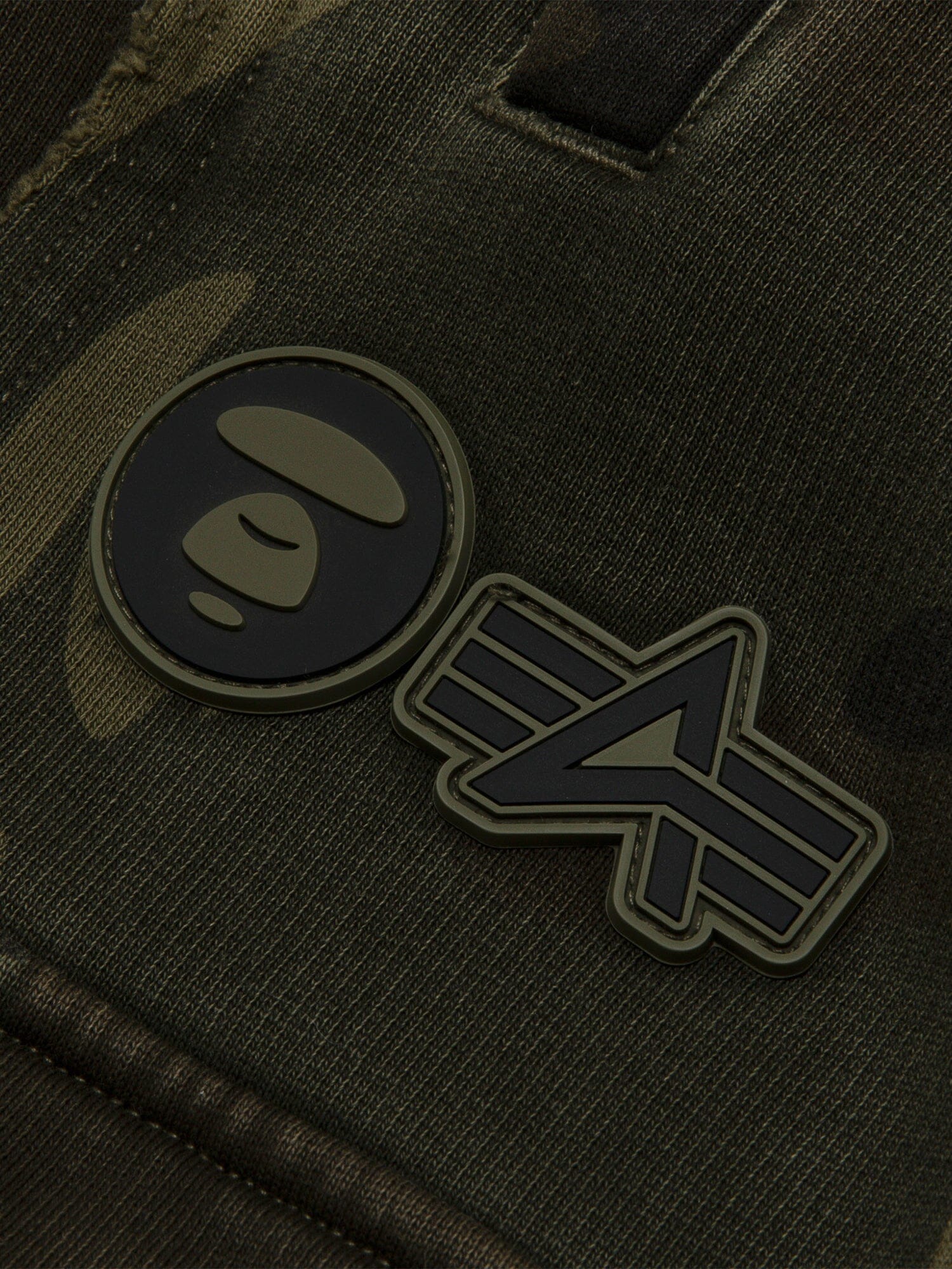 Aape X Alpha Camo Army Sweatpant by Alpha Industries, camo fabric, two rubber patches: circular face and abstract emblem.