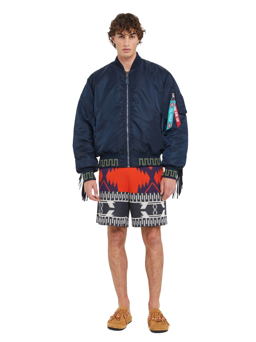 Alanui x Alpha Industries MA-1 jacket with colorful tags and patchwork bandana lining, patterned red, black, and white Alanui shorts, tan moccasins.
