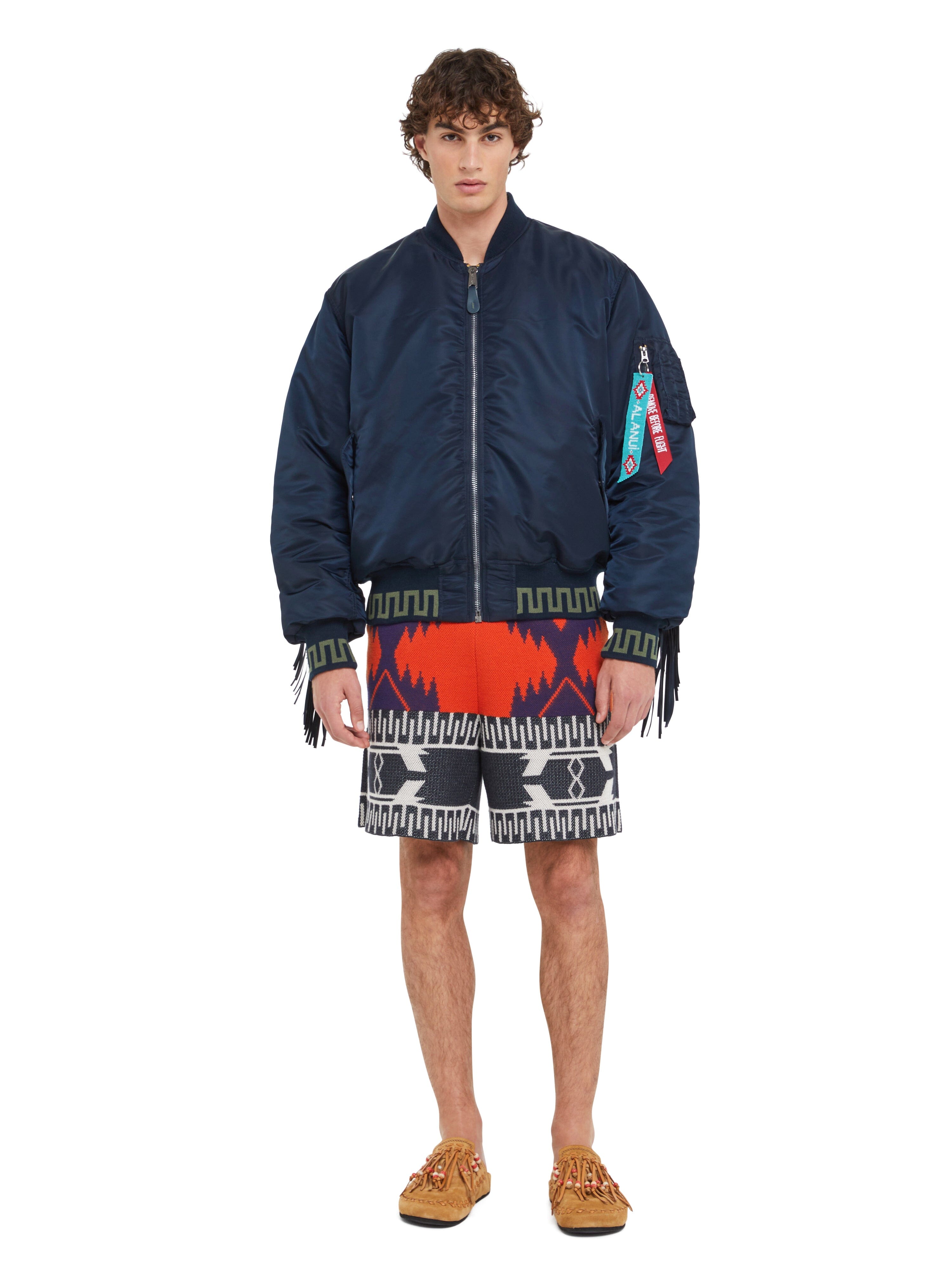 Alanui x Alpha Industries MA-1 jacket with colorful tags and patchwork bandana lining, patterned red, black, and white Alanui shorts, tan moccasins.