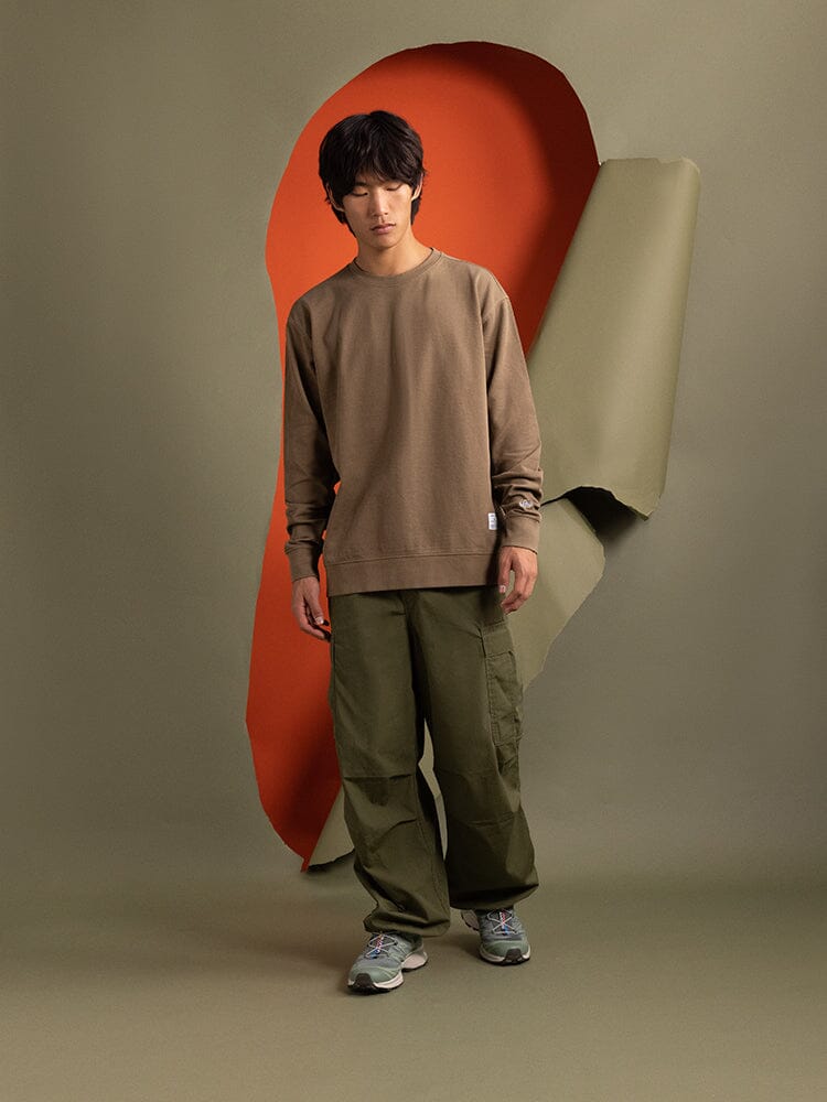 In Front of a Green Wall With an Orange Cutout, a Person Is Wearing the Essential Crewneck From Alpha Industries Along With Green Cargo Pants and Sneakers.