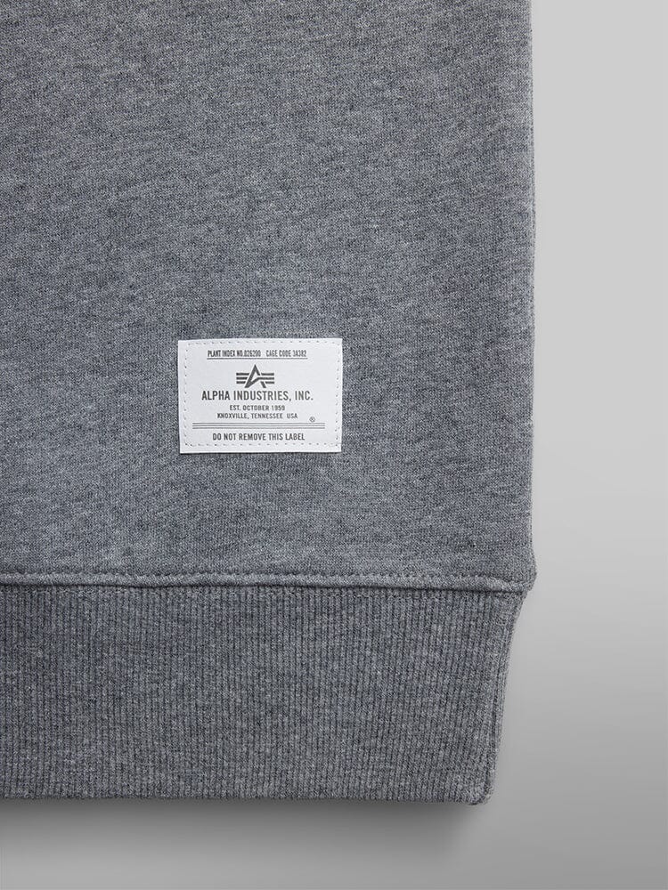 A close-up view of the hem of a gray Essential Crewneck by Alpha Industries showcases its distinctive white logo label, highlighting its refined design and comfortable cotton blend material.