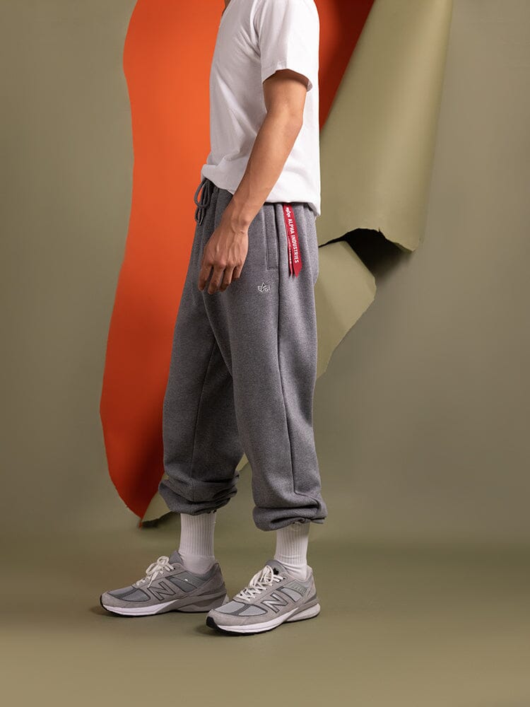 A person wearing a white t-shirt and Essential Sweatpants by Alpha Industries, featuring an elastic waistband, stands sporting comfortable gray New Balance sneakers, perfectly complementing the casual look.