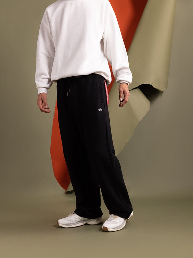 A person wearing a white sweatshirt, Alpha Industries Fw24-Drop1 Essential Sweatpants in black with an elastic waistband, and white sneakers.