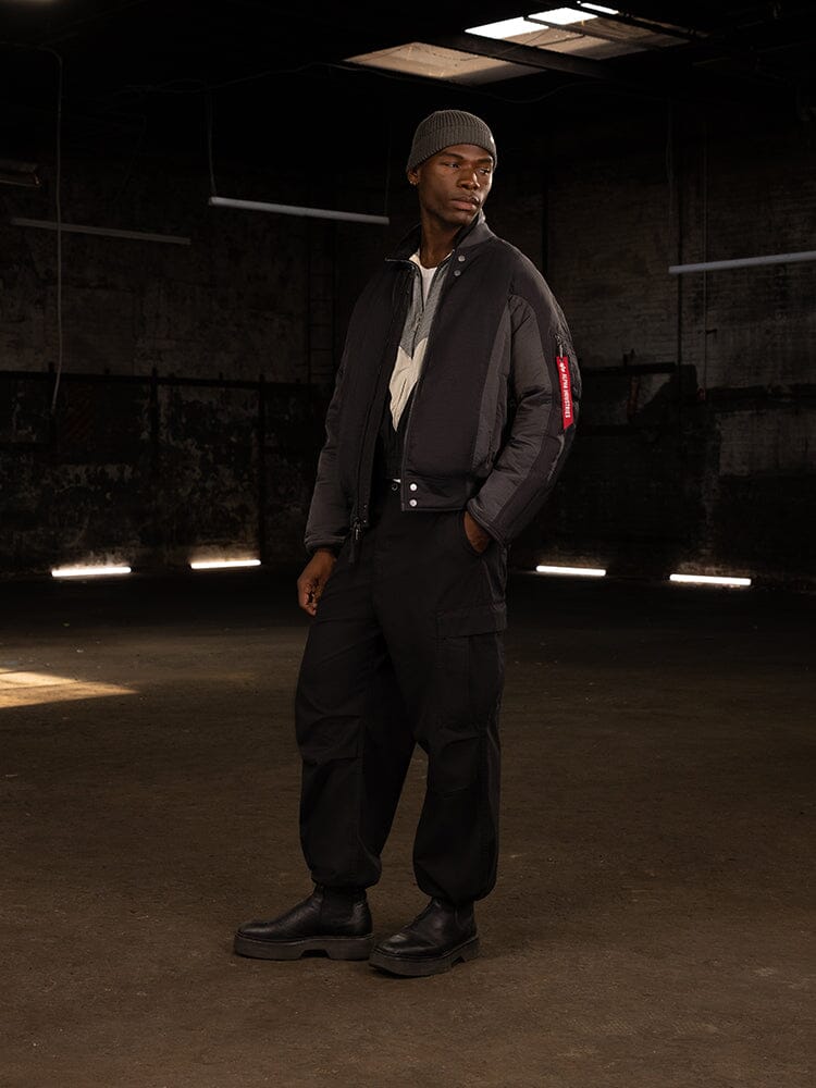 A person stands in a dimly lit industrial space, wearing an Alpha Industries Insulated MA-1 Bomber Jacket, cargo pants, boots, and a beanie.
