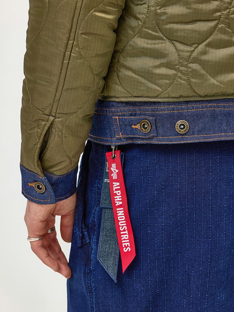 A close-up of an individual wearing the Alpha X Lee Quilted Jacket by Alpha Industries showcases a heritage style featuring green quilted fabric with denim trim. The jacket is adorned with a distinctive red tag from Alpha Industries. This mixed-media fashion ensemble is completed with blue pinstripe pants and a silver ring.