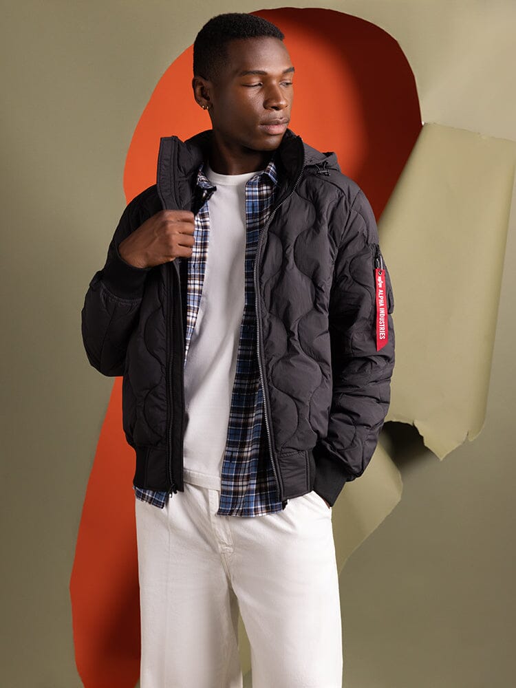 A person wearing the Lightweight Quilted Down Jacket in black from Alpha Industries, featuring a removable hood, over a plaid shirt and white pants.