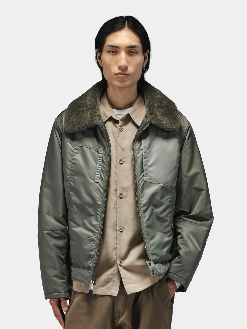 Alpha Industries Nylon Aviator Jacket in olive green with shearling collar, layered over beige button-up shirt and brown pants.