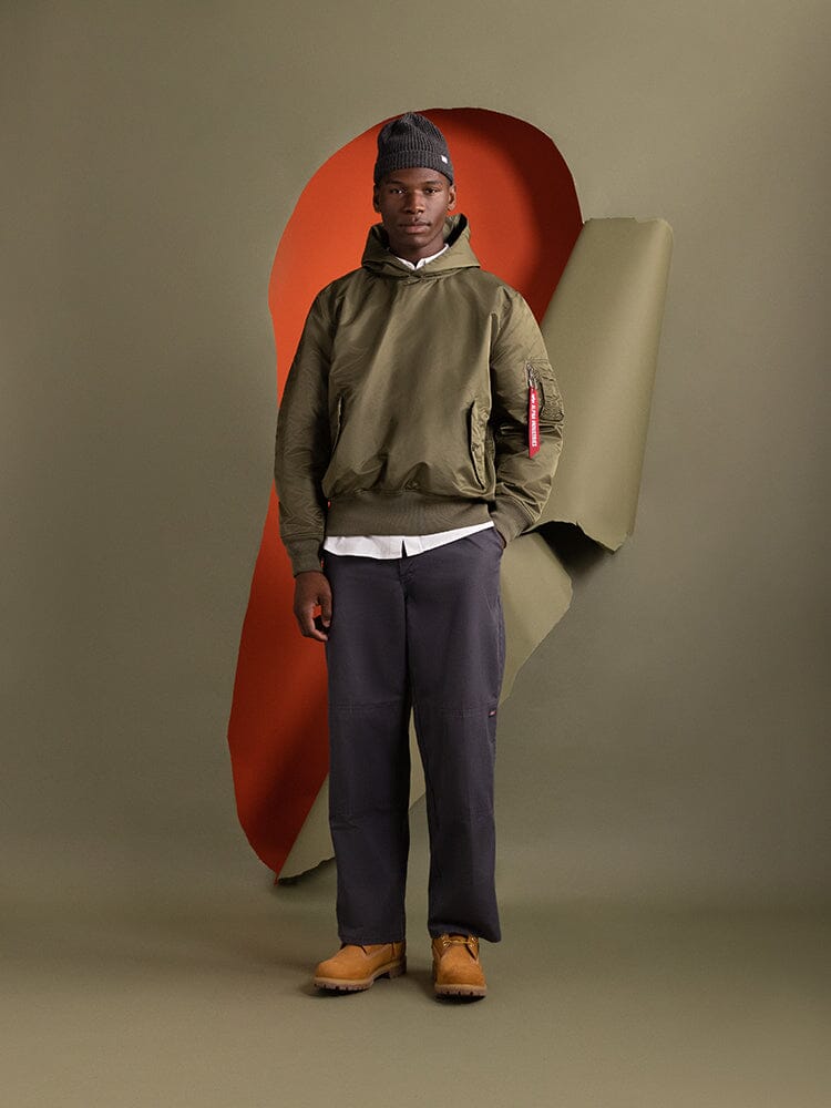 A person wearing the Pull-Over MA-1 Jacket Hoodie by Alpha Industries, with utility pockets in olive green.