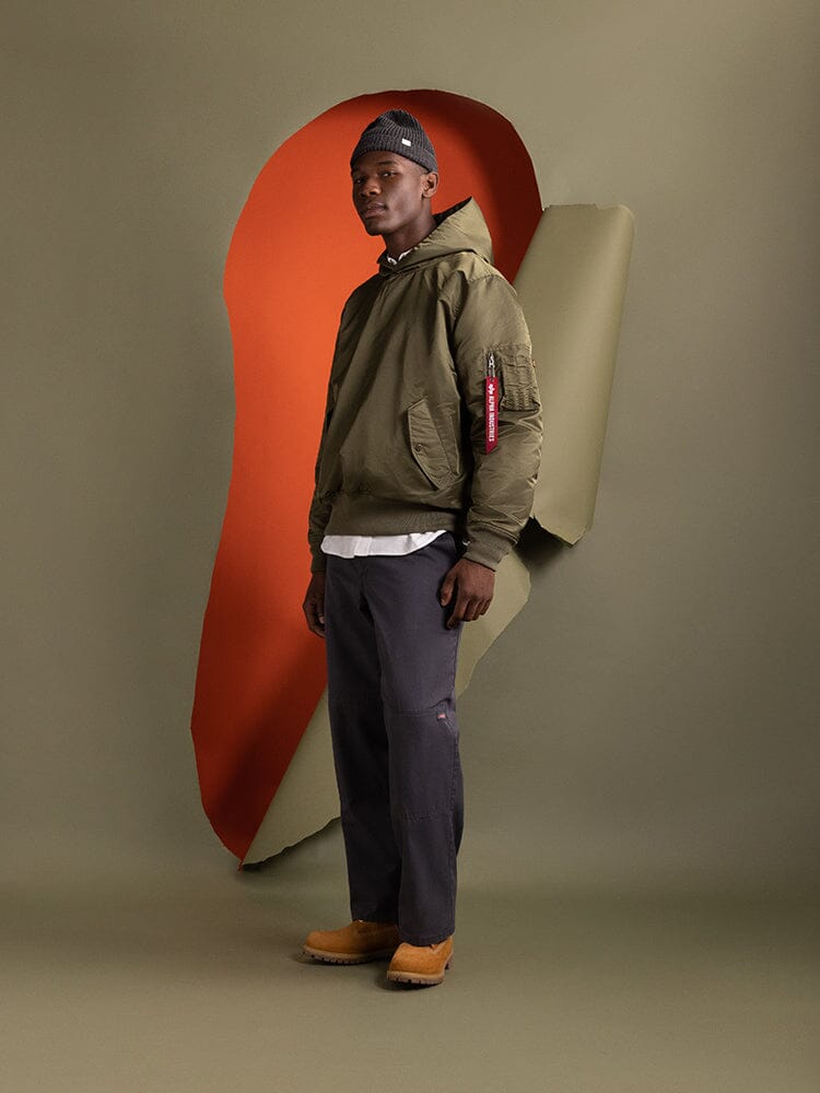 A person stands to the side wearing the Pull-Over MA-1 Jacket Hoodie by Alpha Industries in olive green featuring utility pockets.
