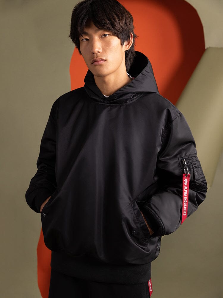 A person dressed in the Pull-Over MA-1 Jacket Hoodie by Alpha Industries, featuring a black design with red accents, highlighting the enduring charm of iconic flight nylon.