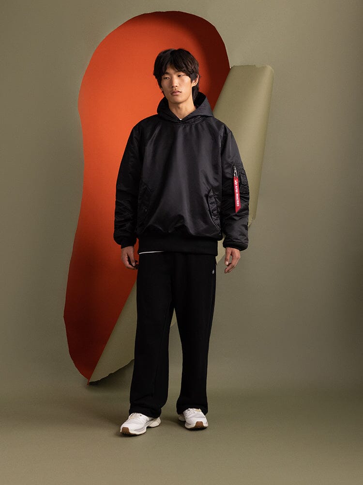 A person is wearing a Pull-Over MA-1 Jacket Hoodie in black by Alpha Industries, paired with black pants featuring utility pockets.