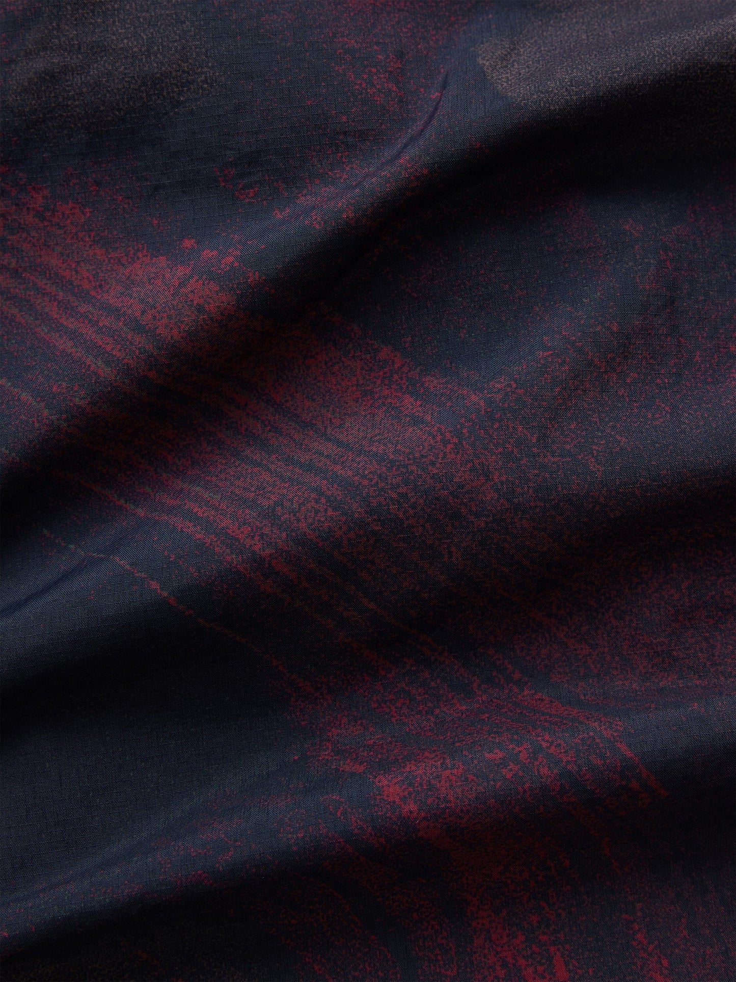 Alpha Industries Reversible M-65 Quilted Liner fabric in dark tones with irregular red patterns and visible folds.