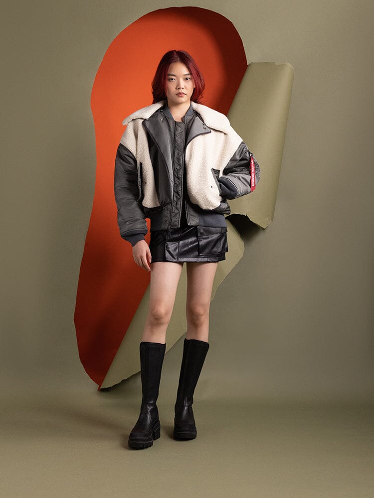 A woman stands wearing the Women's B-15 Sherpa Hybrid Bomber Jacket in field gray by Alpha Industries.