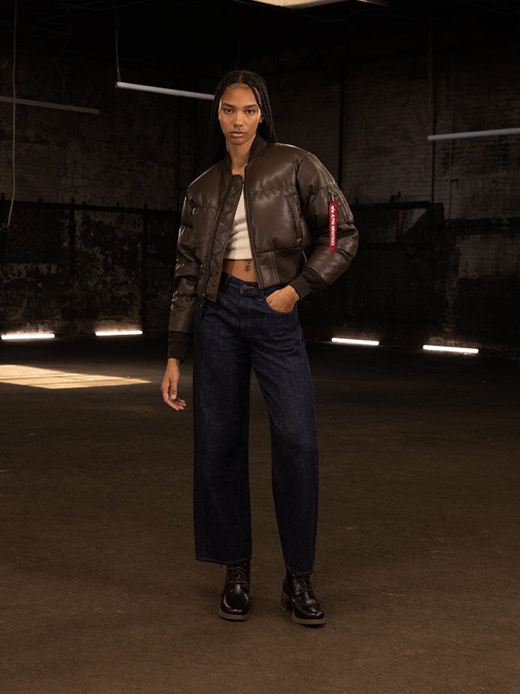 A person wearing the Women's Sierra Faux Leather Short Puffer Jacket by Alpha Industries, paired with jeans, stands in a dimly lit, industrial-looking space.