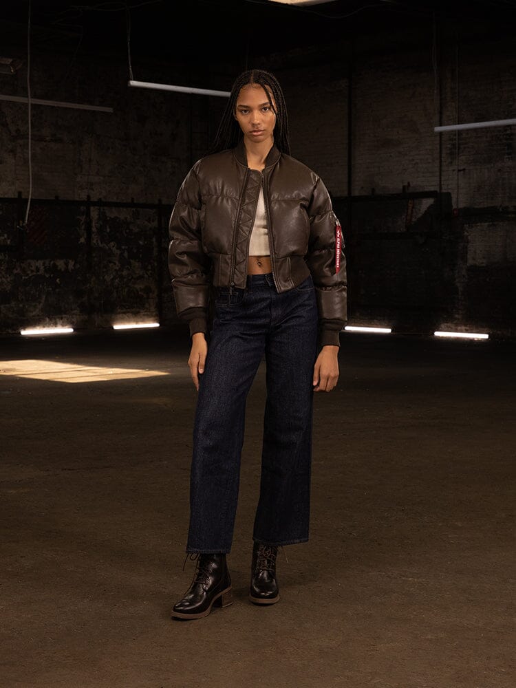 A person stands confidently in their Alpha Industries Women's Sierra Faux Leather Short Puffer Jacket over a white top, paired with dark jeans and black boots.