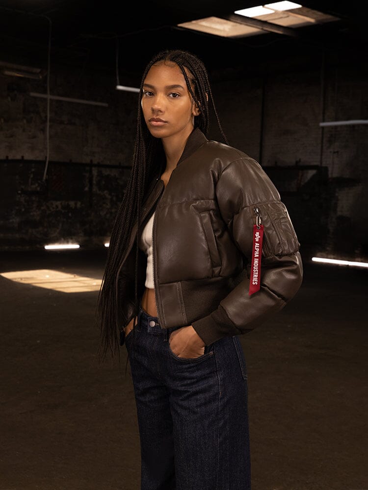 A person wearing the Women's Sierra Faux Leather Short Puffer Jacket from Alpha Industries stands in a dimly lit room, their look enhanced by the rugged elegance of the jacket's cropped fit.