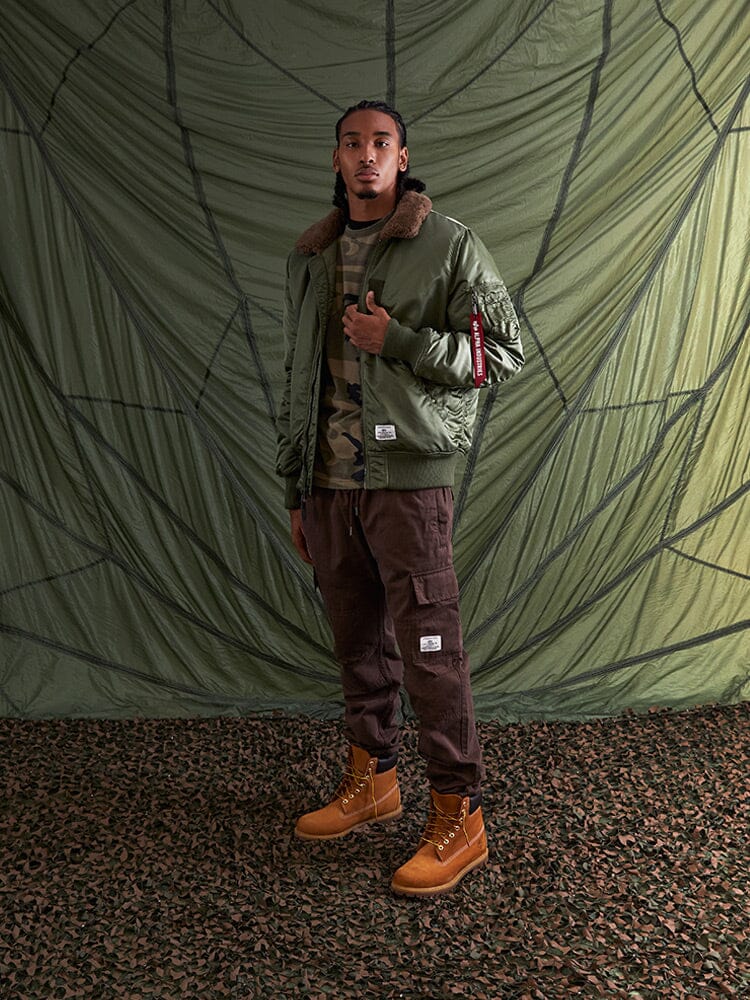 Alpha Industries B-15 Mod Bomber Jacket with faux mouton collar, camo shirt, brown cargo pants, tan boots.