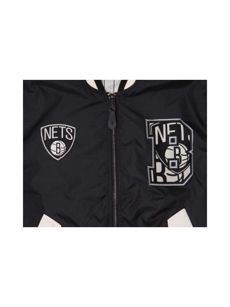 A close-up of the Brooklyn Nets X Alpha X New Era L-2B Bomber Jacket in black, featuring the Brooklyn Nets logo on the left and a stylized NETB design on the right, adorned with team patches and a pinstripe interior.
