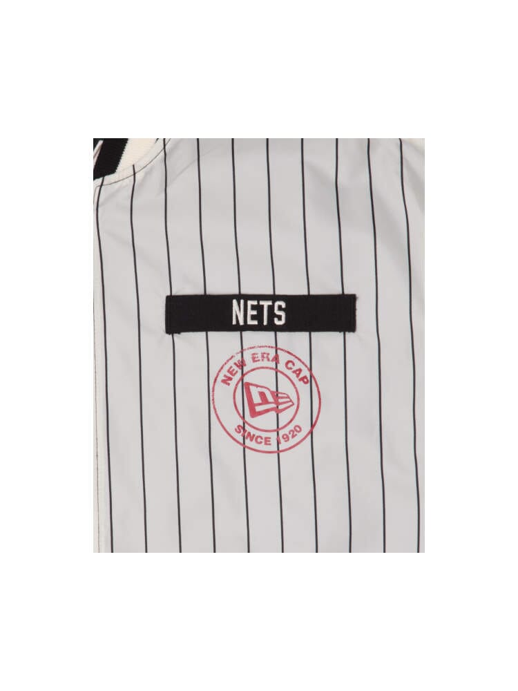 The Brooklyn Nets X Alpha X New Era L-2B Bomber Jacket, made by Alpha Industries, is a gray pinstripe sports jacket adorned with team patches, showcasing a Nets emblem and the New Era Cap Since 1920 logo in a red circle.