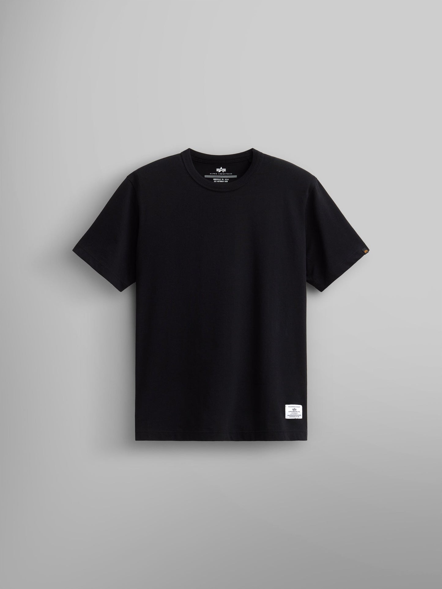Alpha Industries Essential Tee in black cotton with short sleeves, crew neck, and small white label near the bottom hem.