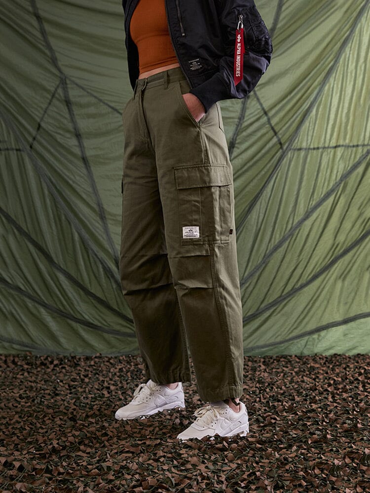 Alpha Industries M-65 Cargo Pant Womens in olive green, white sneakers, orange top, black jacket with red tag.