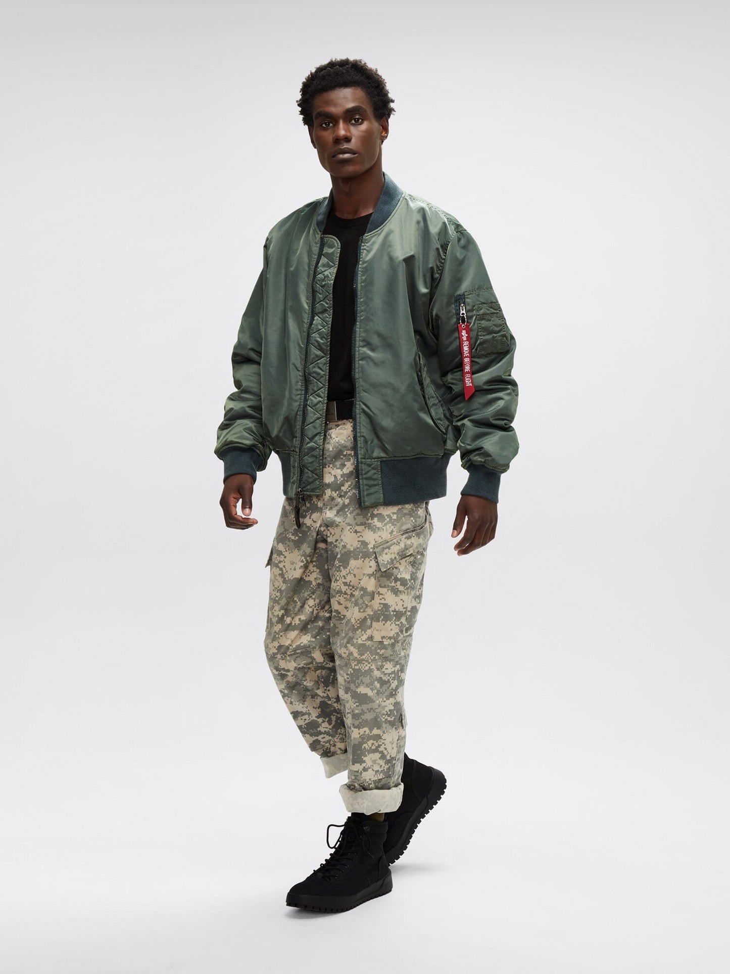 The person dons an MA-1 Battlewash Bomber Jacket by Alpha Industries, Inc., paired with a black shirt, camouflage pants, and rugged black boots, creating a striking look.