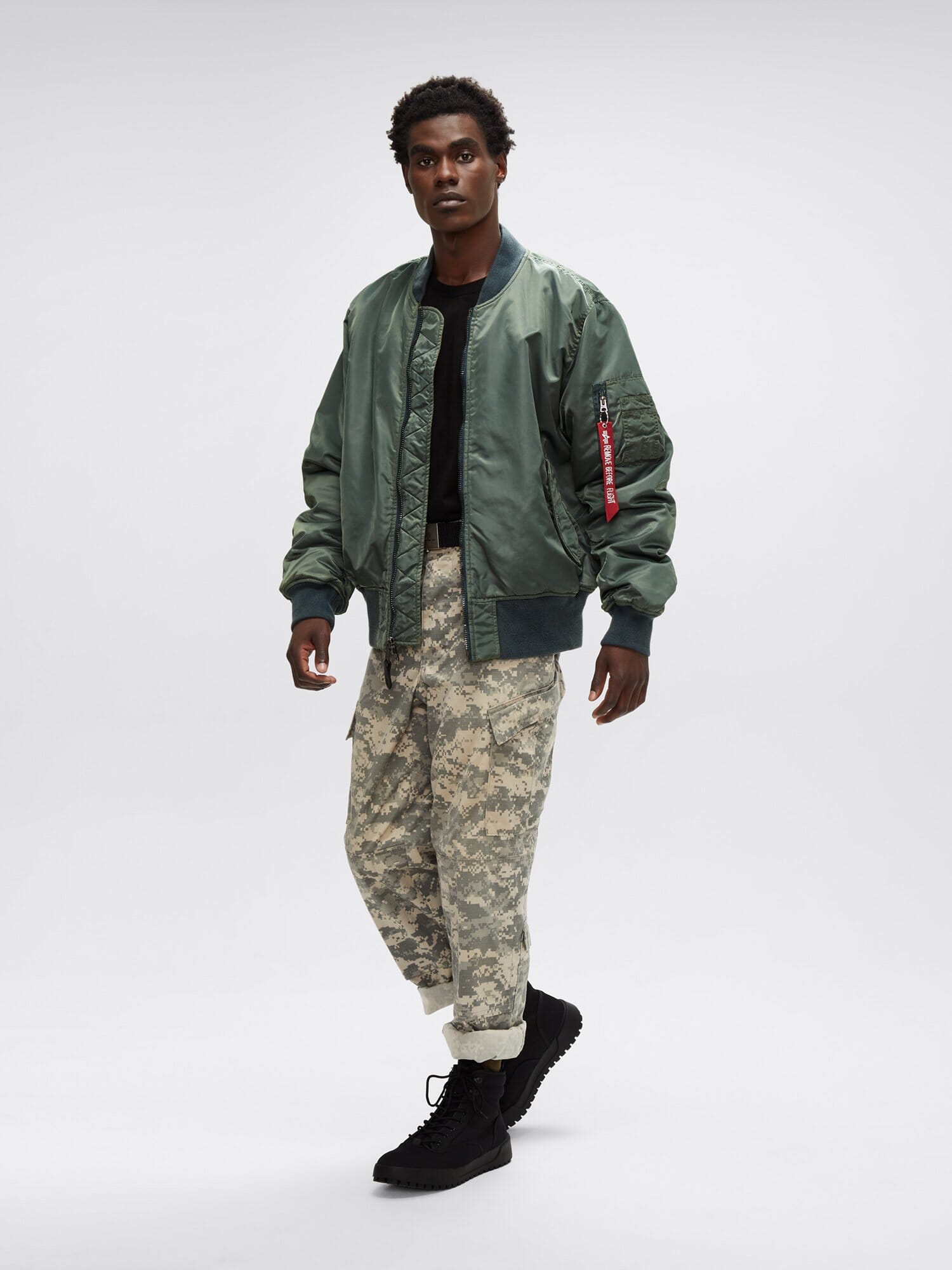 The person dons an MA-1 Battlewash Bomber Jacket by Alpha Industries, Inc., paired with a black shirt, camouflage pants, and rugged black boots, creating a striking look.