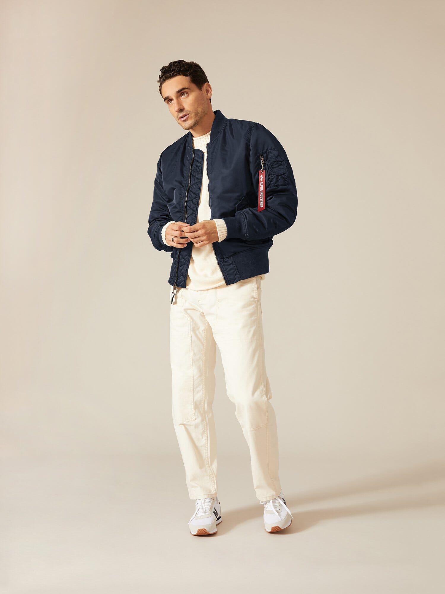 A man is dressed in an Alpha Industries, Inc. MA-1 Battlewash Bomber Jacket in navy, accompanied by a white shirt, white pants, and sneakers.