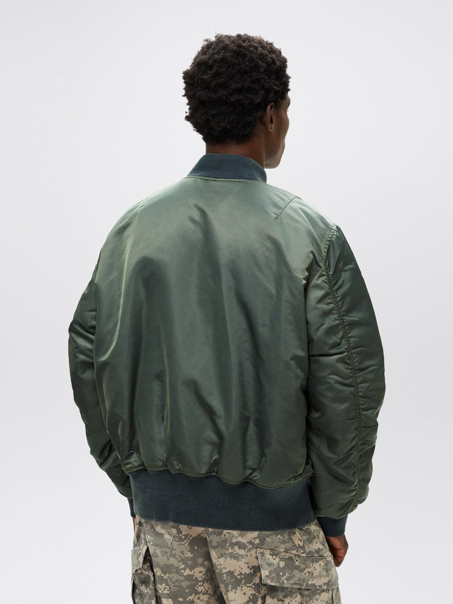 An individual sporting a green MA-1 Battlewash Bomber Jacket from Alpha Industries and camo pants, positioned with their back to the camera. The battlewash finish of the jacket enhances the outfit with a rugged charm.