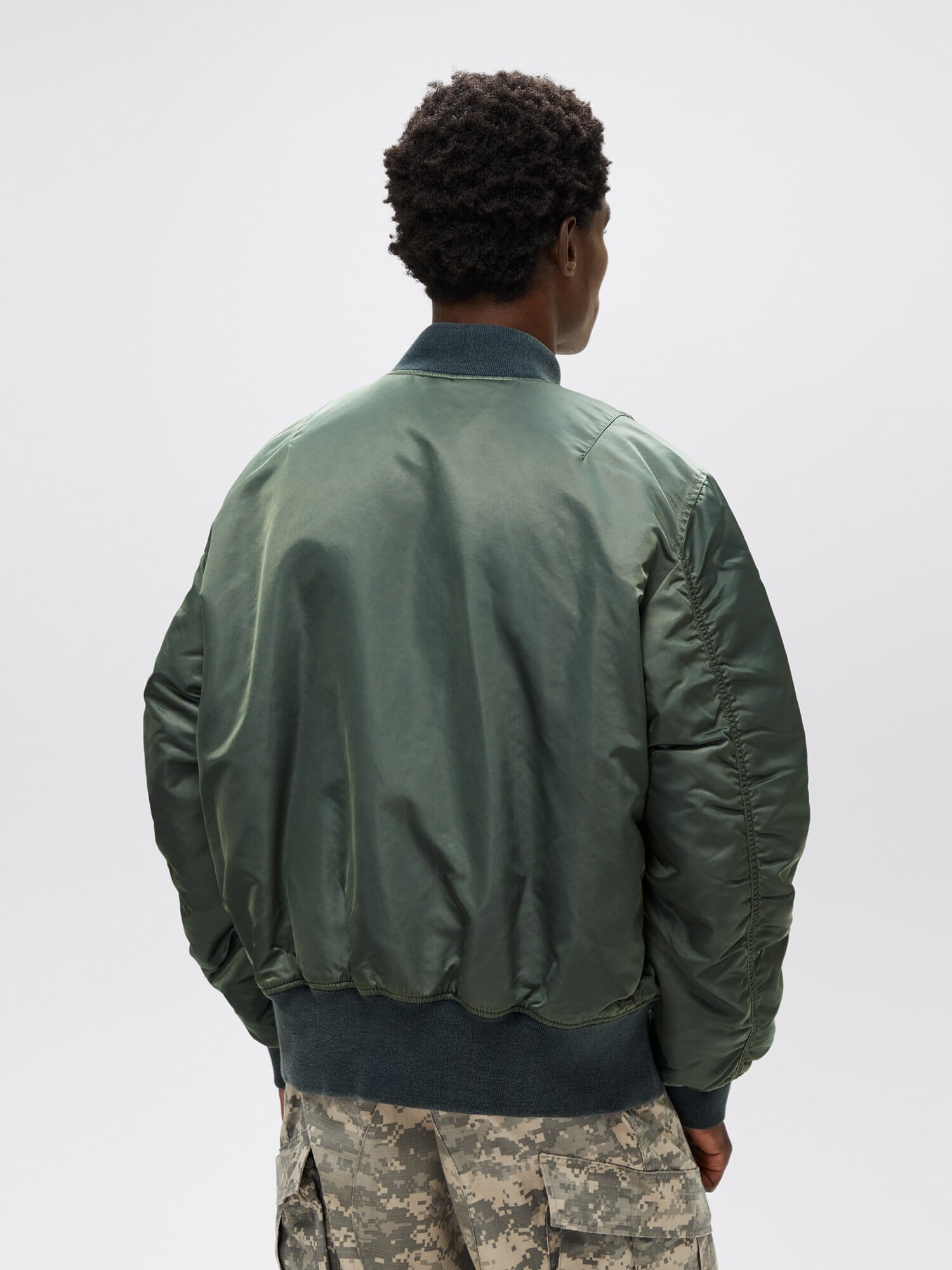 An individual sporting a green MA-1 Battlewash Bomber Jacket from Alpha Industries and camo pants, positioned with their back to the camera. The battlewash finish of the jacket enhances the outfit with a rugged charm.