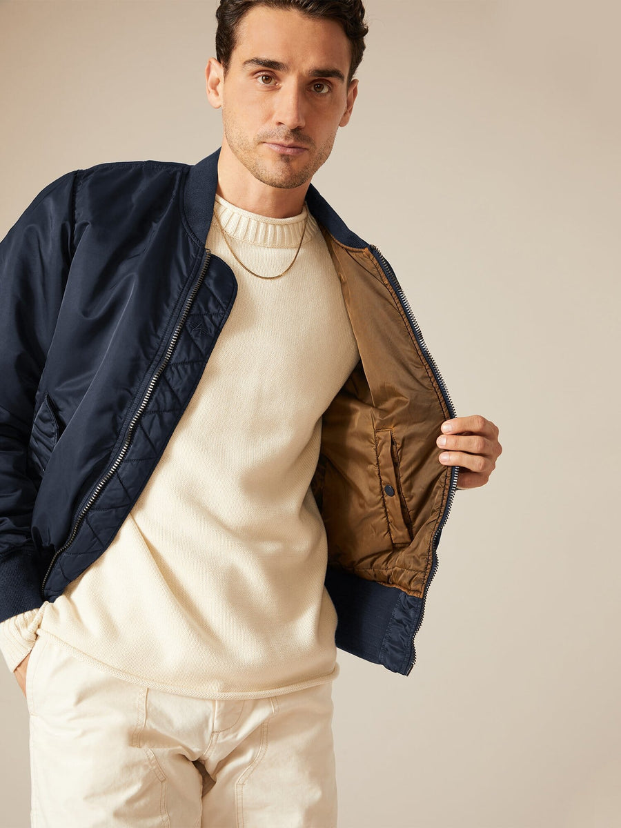 A man wearing a cream sweater and cream pants subtly reveals the brown lining of his navy MA-1 Battlewash Bomber Jacket by Alpha Industries.