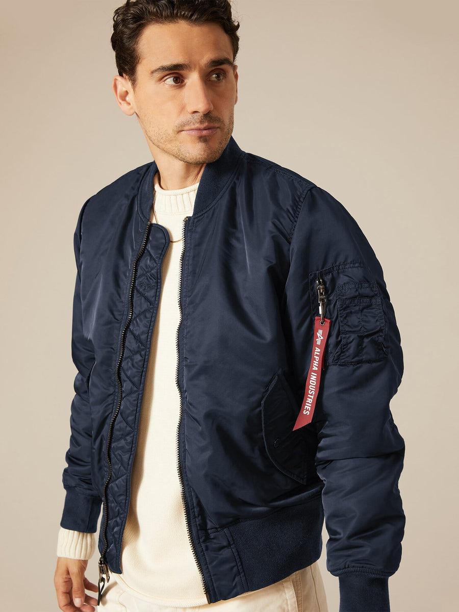 A man wearing a navy MA-1 Battlewash Bomber Jacket by Alpha Industries, Inc., featuring a distinctive red tag on the sleeve, pairs it with a crisp white shirt as he looks to the side.