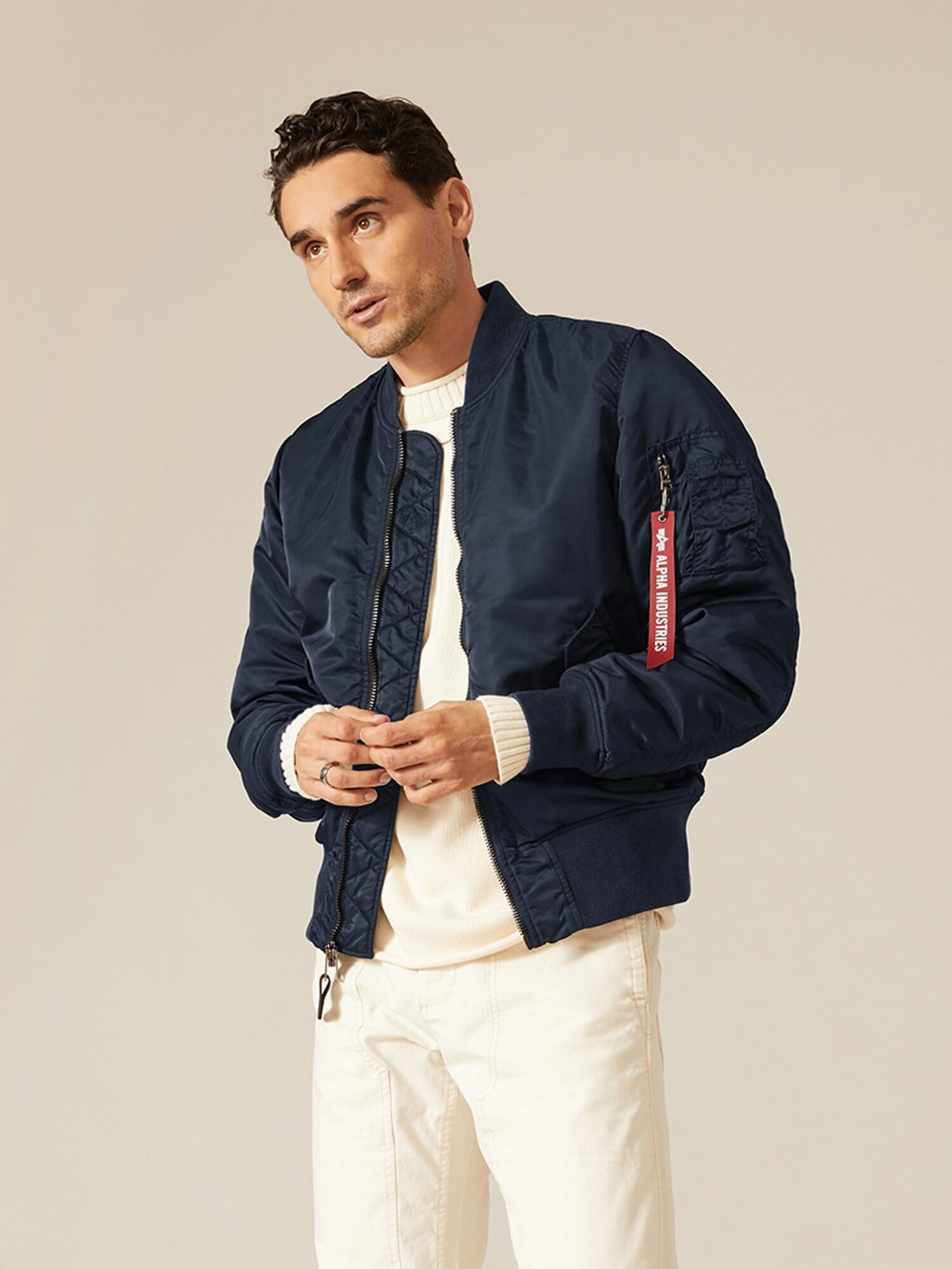 A person wearing an Alpha Industries, Inc. MA-1 Battlewash Bomber Jacket in navy over a white sweater and pants.