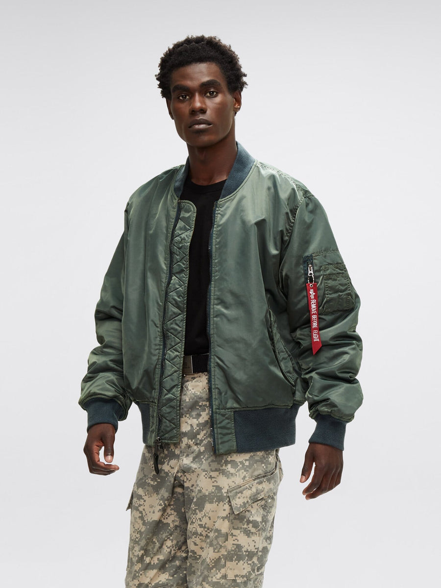 A person wearing an Alpha Industries MA-1 Battlewash Bomber Jacket in green, paired with a black shirt and battlewash camouflage pants.