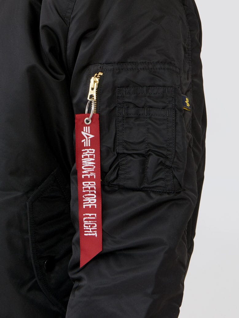 Close-up of sleeve pocket on black Alpha Industries MA-1 Bomber Jacket Slim Fit showing red “Remove Before Flight” tag and gold zipper.