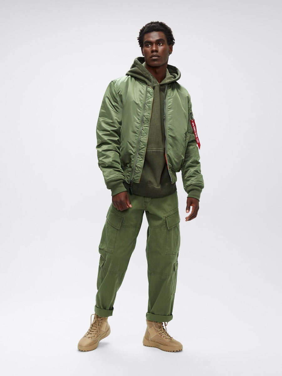 Three-quarter full-body front view of sage green Alpha Industries MA-1 Bomber Jacket Slim Fit showing ribbed cuffs, waistband, and red sleeve tag.