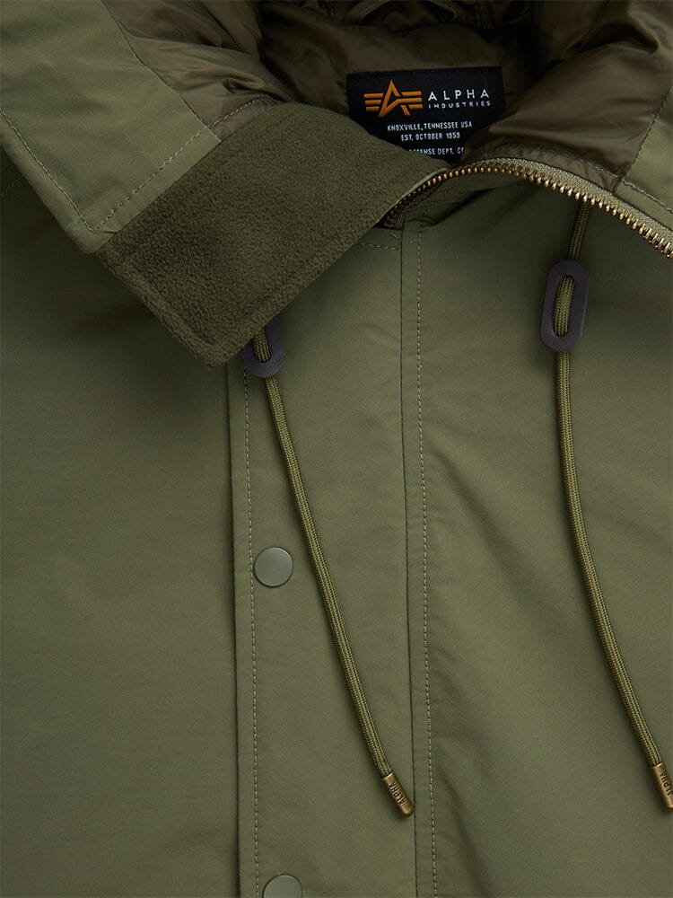 Close-up of an olive green N-2B Altitude Parka highlighting its collar, zipper, drawstrings, and snap buttons. The inside label displays Alpha Industries with care instructions.