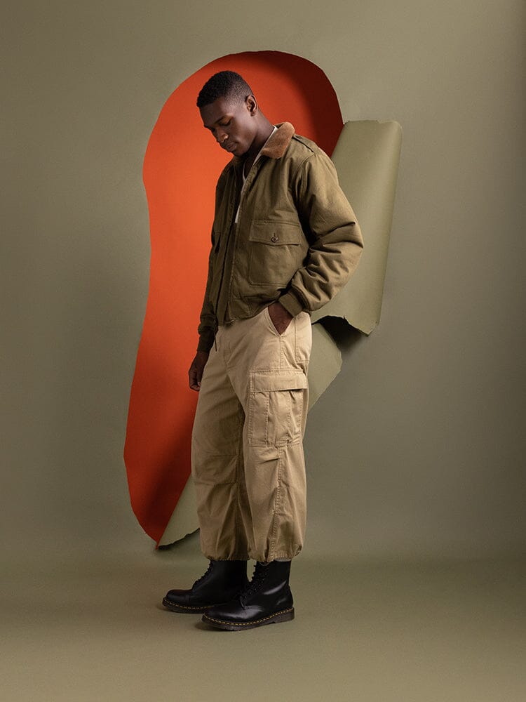 A man stands wearing a Retro-Style 1940s B-10 Bomber Jacket in olive by Alpha Industries, paired with beige cargo pants and black boots.
