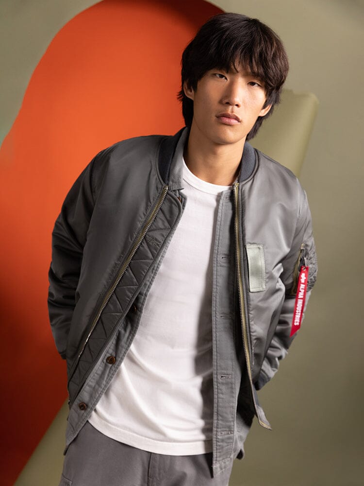 A person wearing an Alpha Industries 1960s MA-1 Bomber Jacket in gray and a white T-shirt.