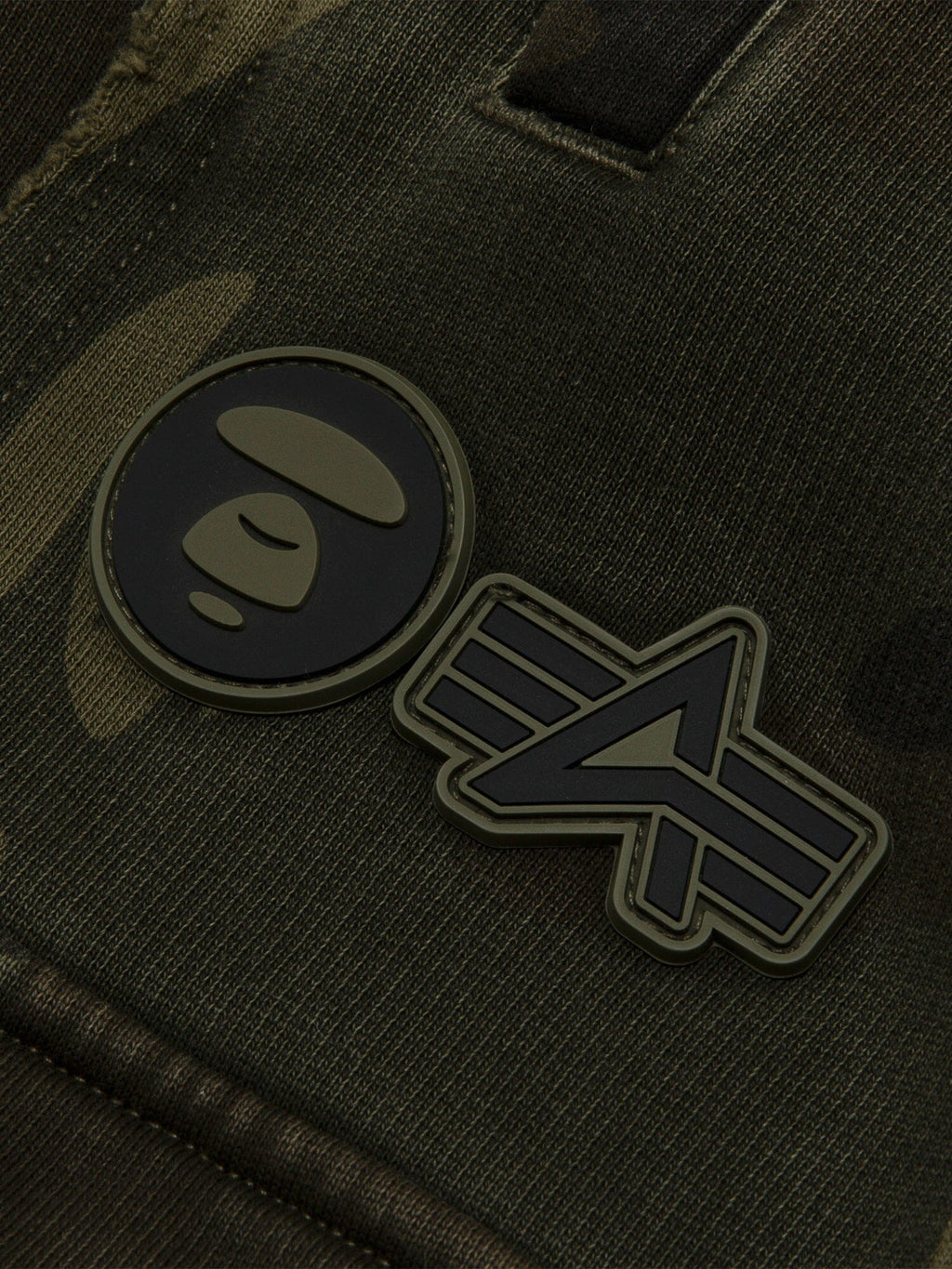 Aape X Alpha Camo Army Sweatpant by Alpha Industries, camo fabric, two rubber patches: circular face and abstract emblem.