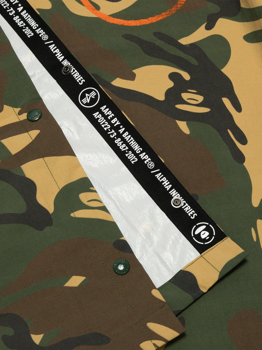Close-up of AAPE X Alpha Industries 2 Layer Rain Jacket With Camouflaged Fabric In Green And Brown Tones, White Lining, And Black And White Strap With Aape By *A Bathing Ape® And Alpha Industries.
