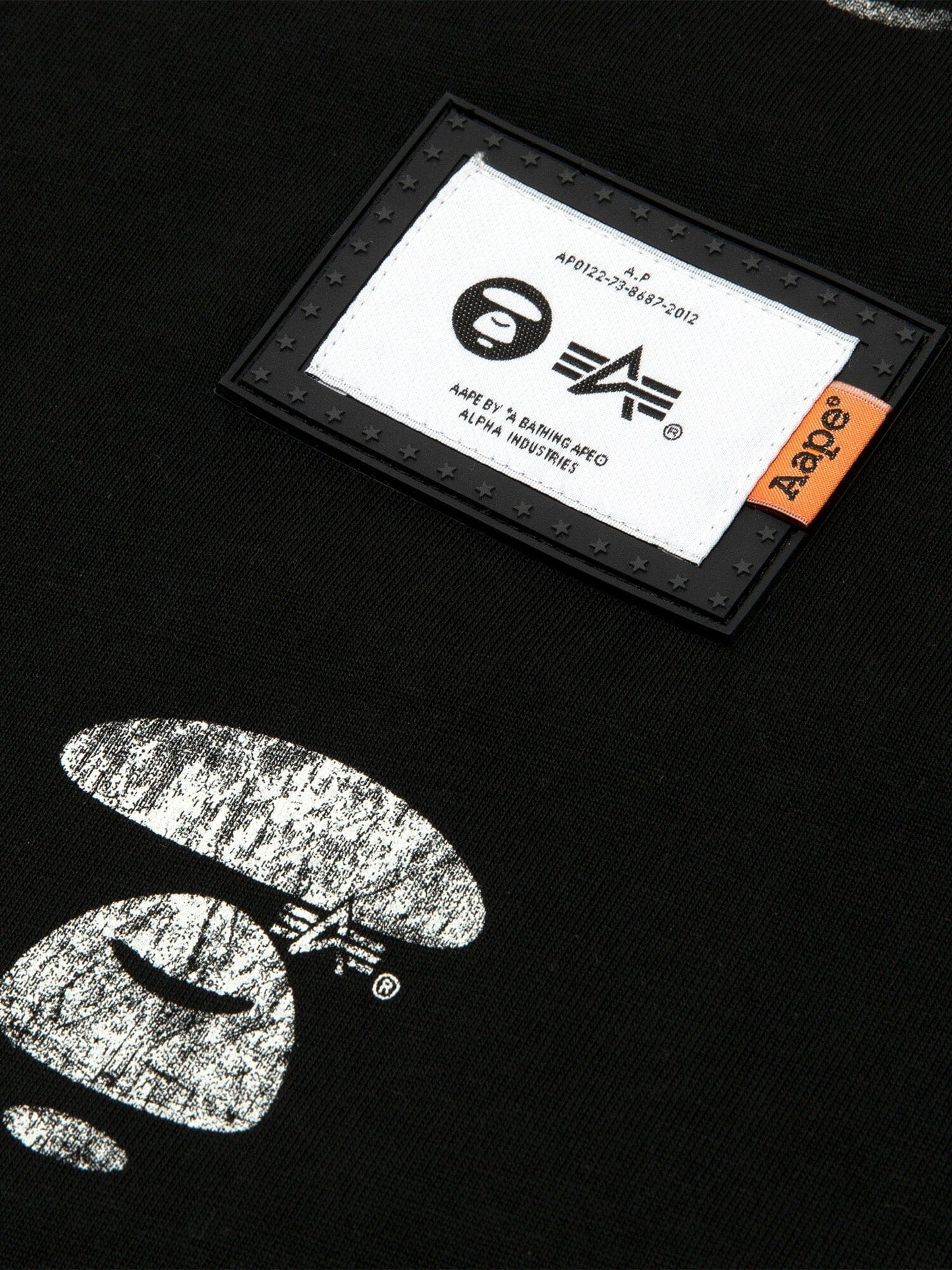 Close-up of the Black AAPE x Alpha Industries Somewhere in Universe Tee with Vintage Distressed Look, AAPE Logo Patch, White Graphics, Small Orange Tag.